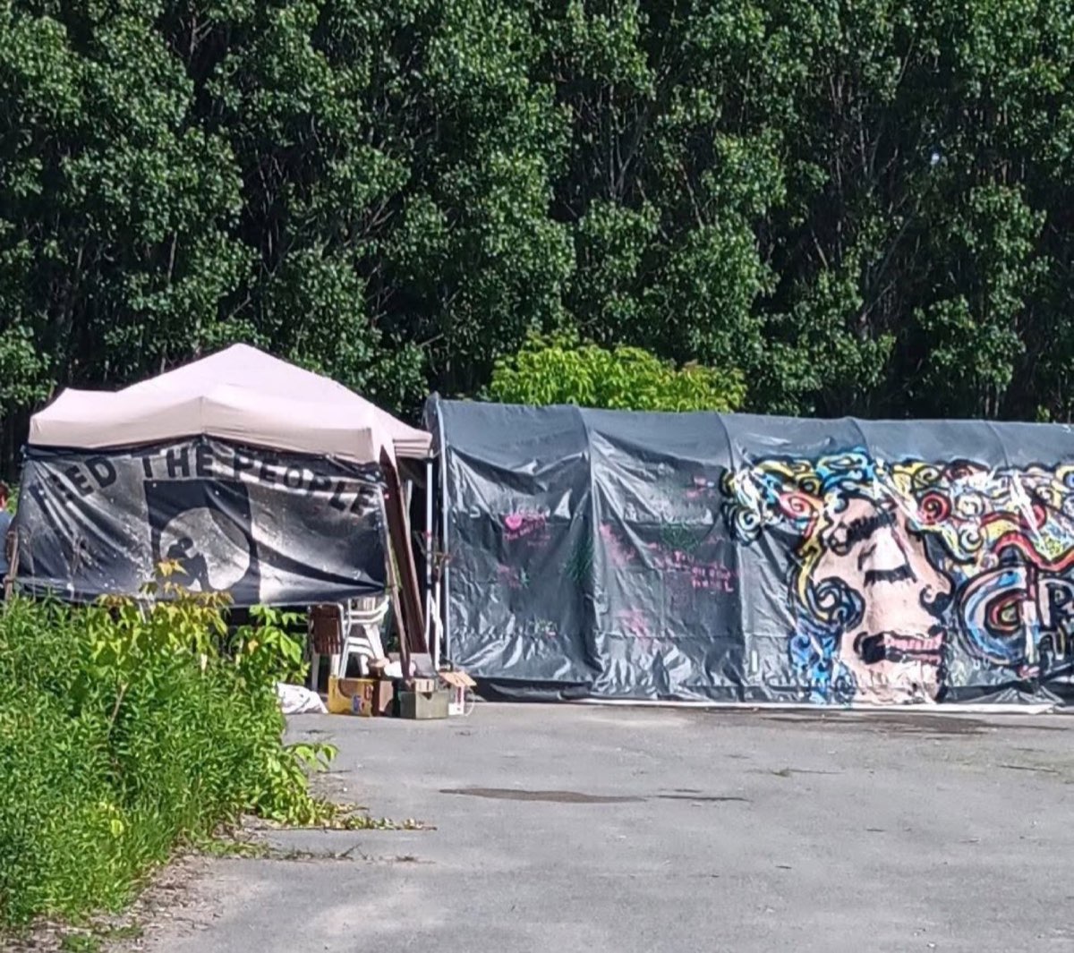 The Feed the People pop-up canopy and supply tent set up on the gravel lot at Belle Park, with trees behind