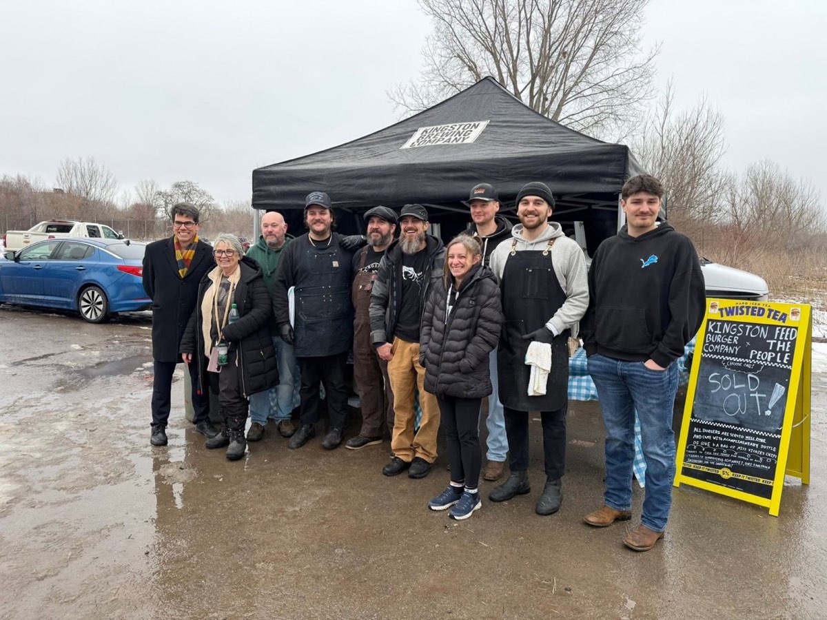 A crew of Feed the People volunteers posing outside the Kingston Brewing Company tent behind a sold-out burger day sandwich board