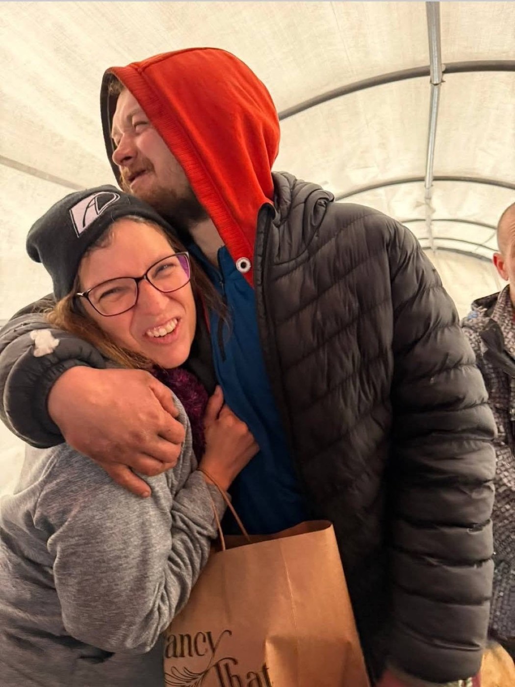 Two friends embrace inside the Belle Park tent, one holding a paper bag of food