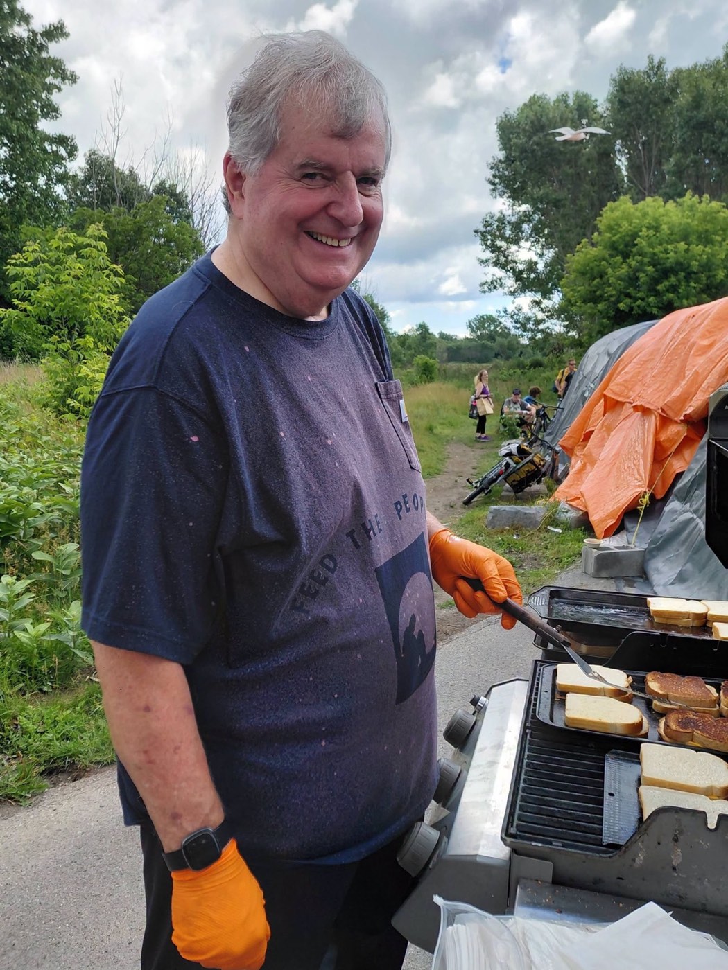 A volunteer in a Feed the People shirt grills sandwiches on an outdoor propane grill beside the tent at Belle Park