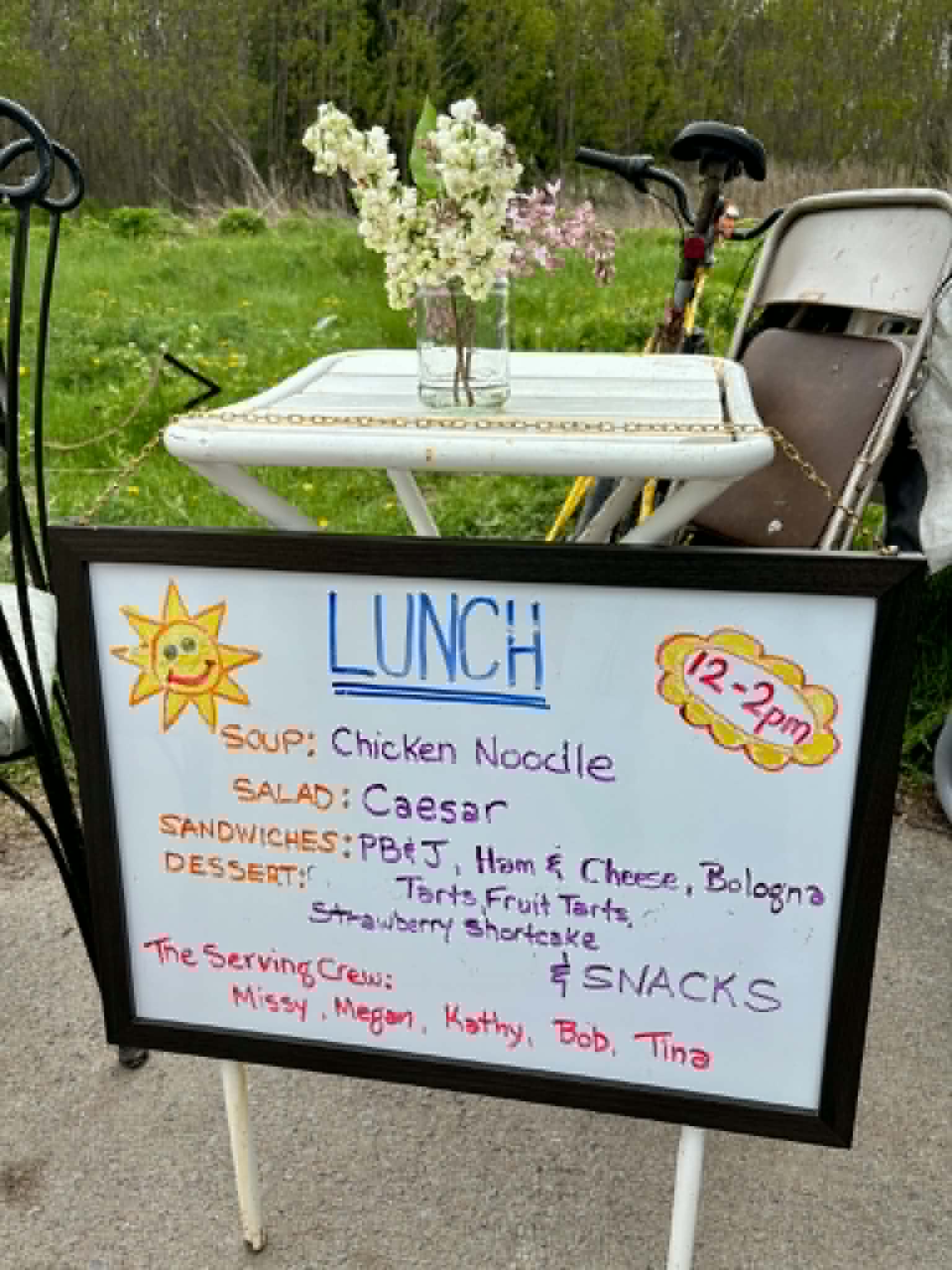 The hand-drawn LUNCH menu whiteboard at Belle Park listing chicken noodle soup, Caesar salad, sandwiches and desserts, with lilacs on the table behind it