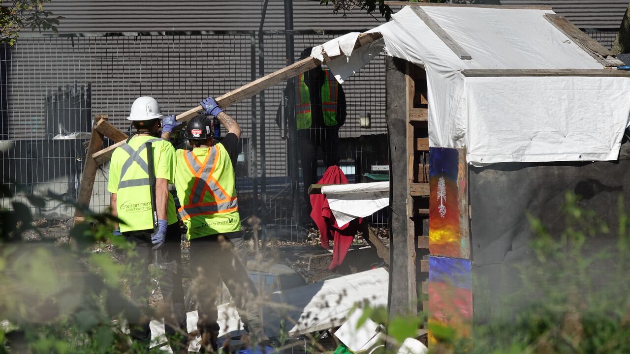Two workers in high-visibility vests and hard hats pull boards from a makeshift shelter at the Belle Park encampment. Photo: Dan Taekema/CBC
