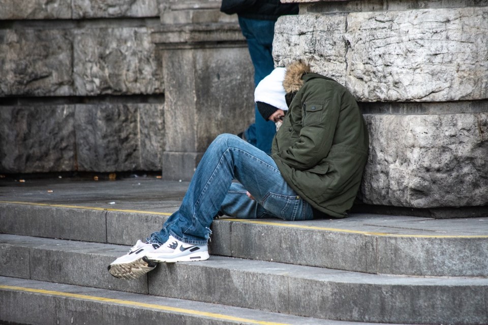 A person in a winter parka and toque sits hunched on cold stone steps beside a building