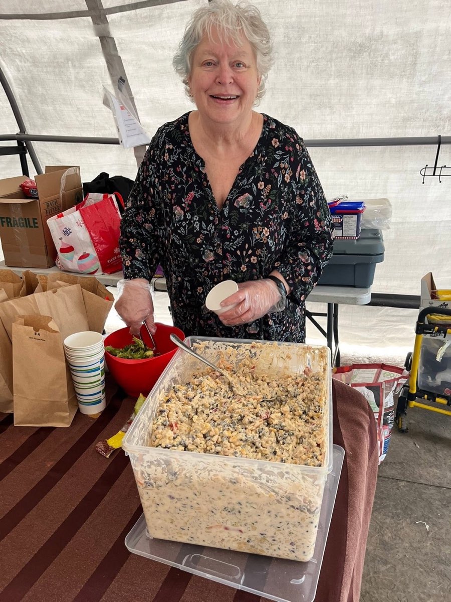 A smiling volunteer scoops from a large tub of salad at the serving table inside the Belle Park tent