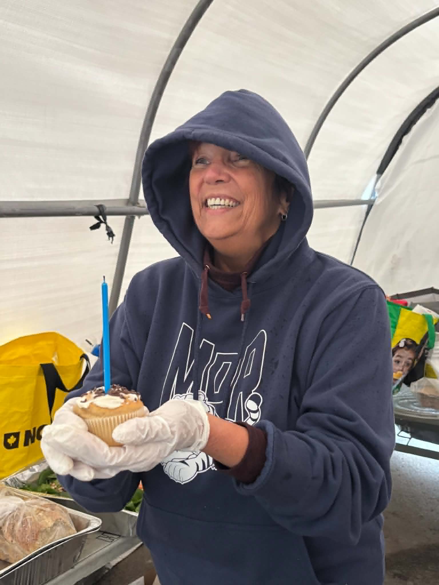 A volunteer in a hooded sweatshirt laughing while holding a cupcake with a single blue birthday candle, inside the Belle Park tent