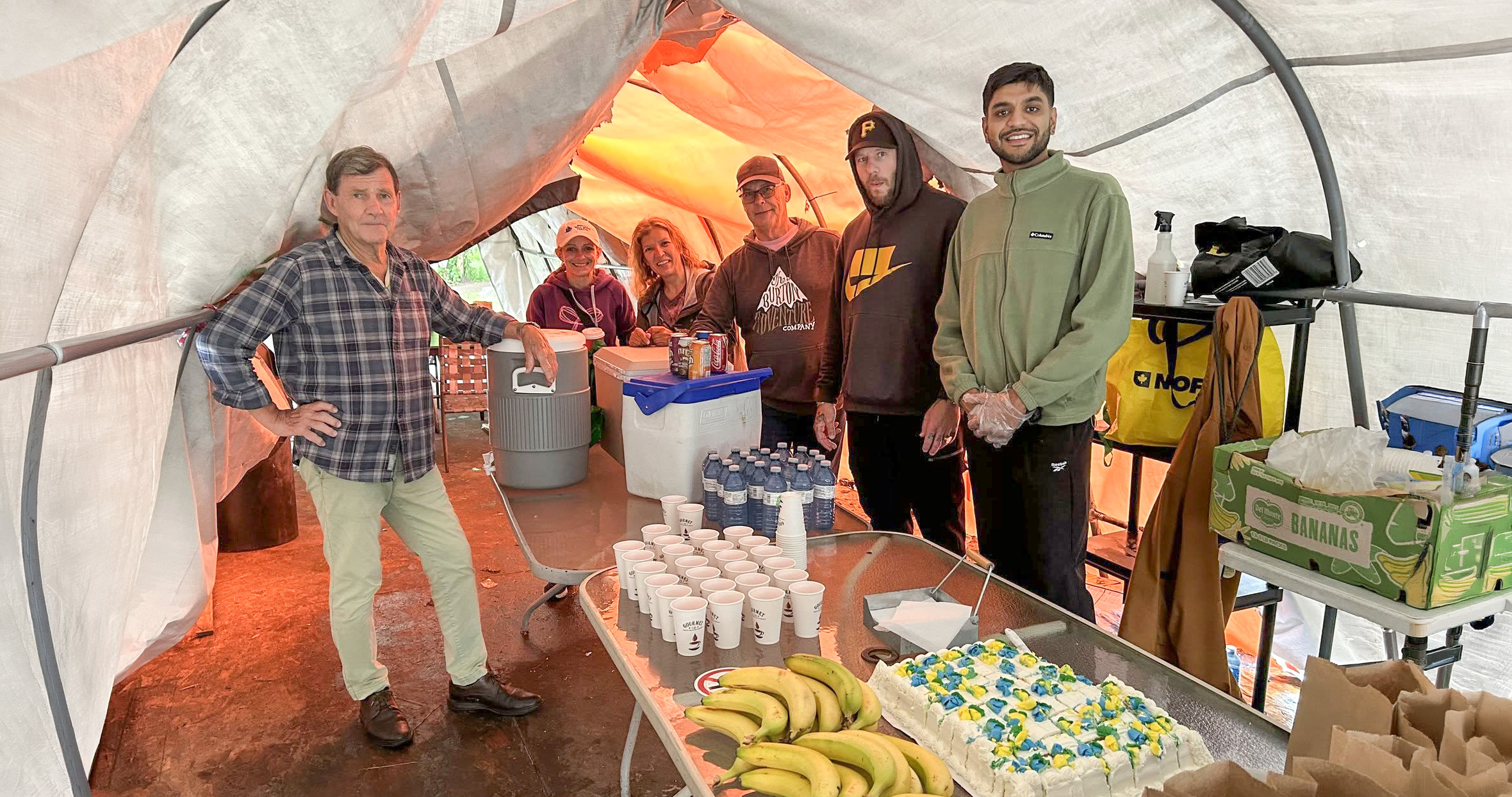 Six Feed the People volunteers beside tables of drinks, bananas and cake inside the tent at Belle Park.