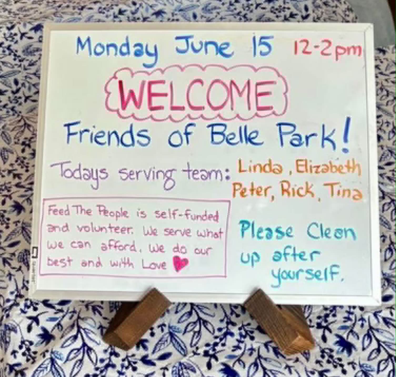 A hand-lettered welcome board at Belle Park naming the day's serving team, with a note reading: Feed the People is self-funded and volunteer. We serve what we can afford, we do our best and with love.