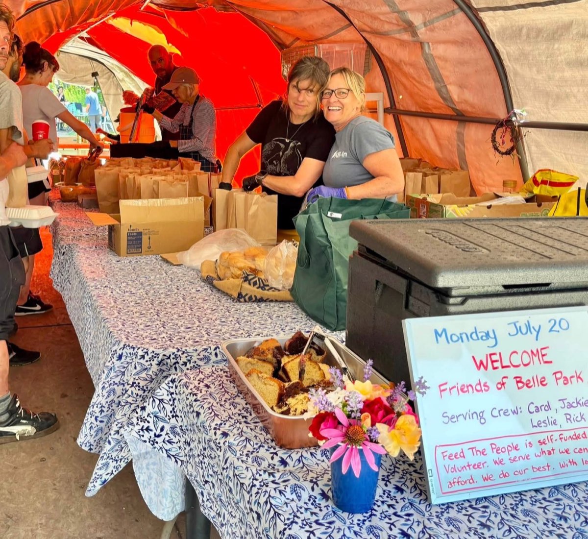 Two volunteers embrace at the Feed the People welcome table at Belle Park, beside trays of prepared food and a handwritten welcome sign